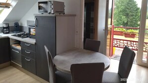 A compact kitchen with a built-in oven, a coffee maker, and a small refrigerator. There is a dining table with chairs and a view of a balcony with a railing and greenery outside. - La Chèvrerie (Taintrux)