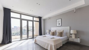 Quarto moderno com uma cama grande, mesa de cabeceira, abajur e vista da cidade através de janelas amplas.