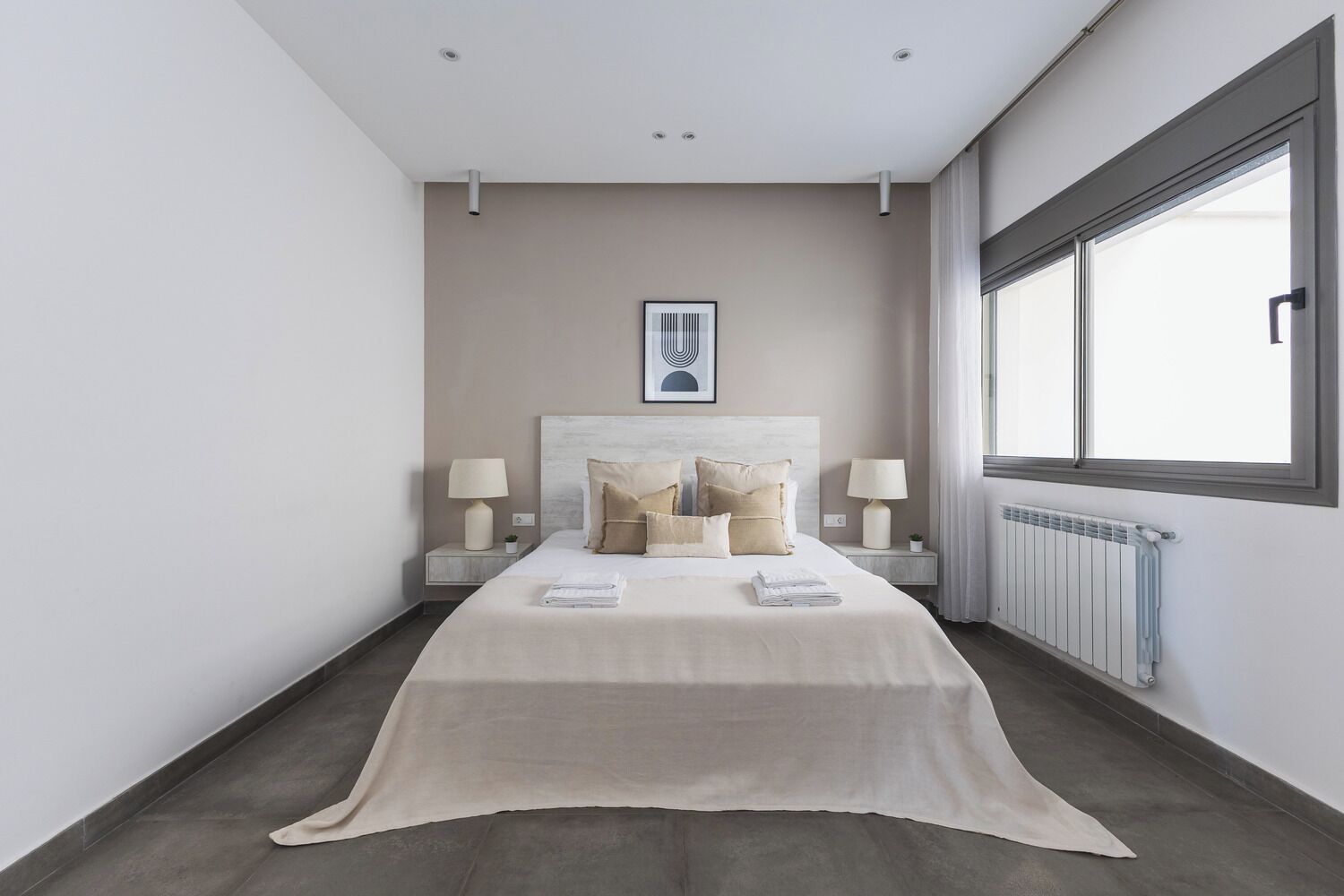 Een moderne hotelkamer met een groot bed, twee nachtkastjes met lampen, een radiator en een groot raam.