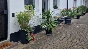 Une rangée de bâtiments blancs aux fenêtres à encadrement noir et dotés d’un toit en tuiles. Devant chaque bâtiment, on trouve des plantes en pot.