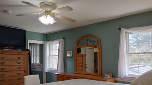 A bedroom with a bed, dresser, vanity, ceiling fan, and large windows.