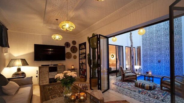 A modern living room with a flat-screen TV, a coffee table with flowers, and a large window opening to a blue-tiled room. - Riad Familial Privatisé au Calme Avec vue sur Marrakech (Marrakech)