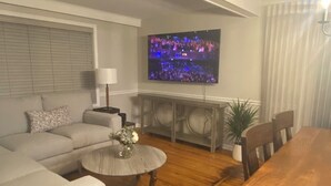 Una sala de estar con sofá, mesa de centro, televisor montado en la pared y una mesa de comedor con sillas.