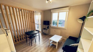 Une pièce avec un plancher en bois, une table à manger avec des chaises, une table basse blanche, un canapé bleu, un meuble blanc, un téléviseur et une fenêtre donnant sur l’extérieur.