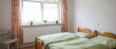 A bedroom with two beds, a window with curtains, a radiator, a chair, and a hanging light fixture.