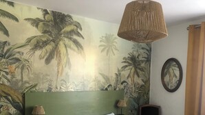 Un dormitorio con cama, mesita de noche, lámpara, silla y una pared con un mural tropical.