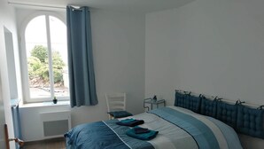 A bedroom with a large bed, a window with curtains, a chair, and a small table with a plant. - Gîte du Haut Montbrault (Fleurigné)