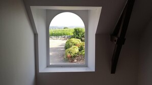 A view from a window with an arched frame, looking out to a paved area with greenery and a distant landscape. - Gîte du Haut Montbrault (Fleurigné)