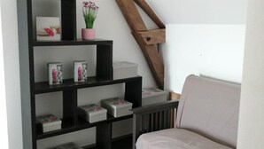 A cozy room with a wooden sloped ceiling, a black shelving unit with decorative items, and a light-colored sofa. - Gîte du Haut Montbrault (Fleurigné)
