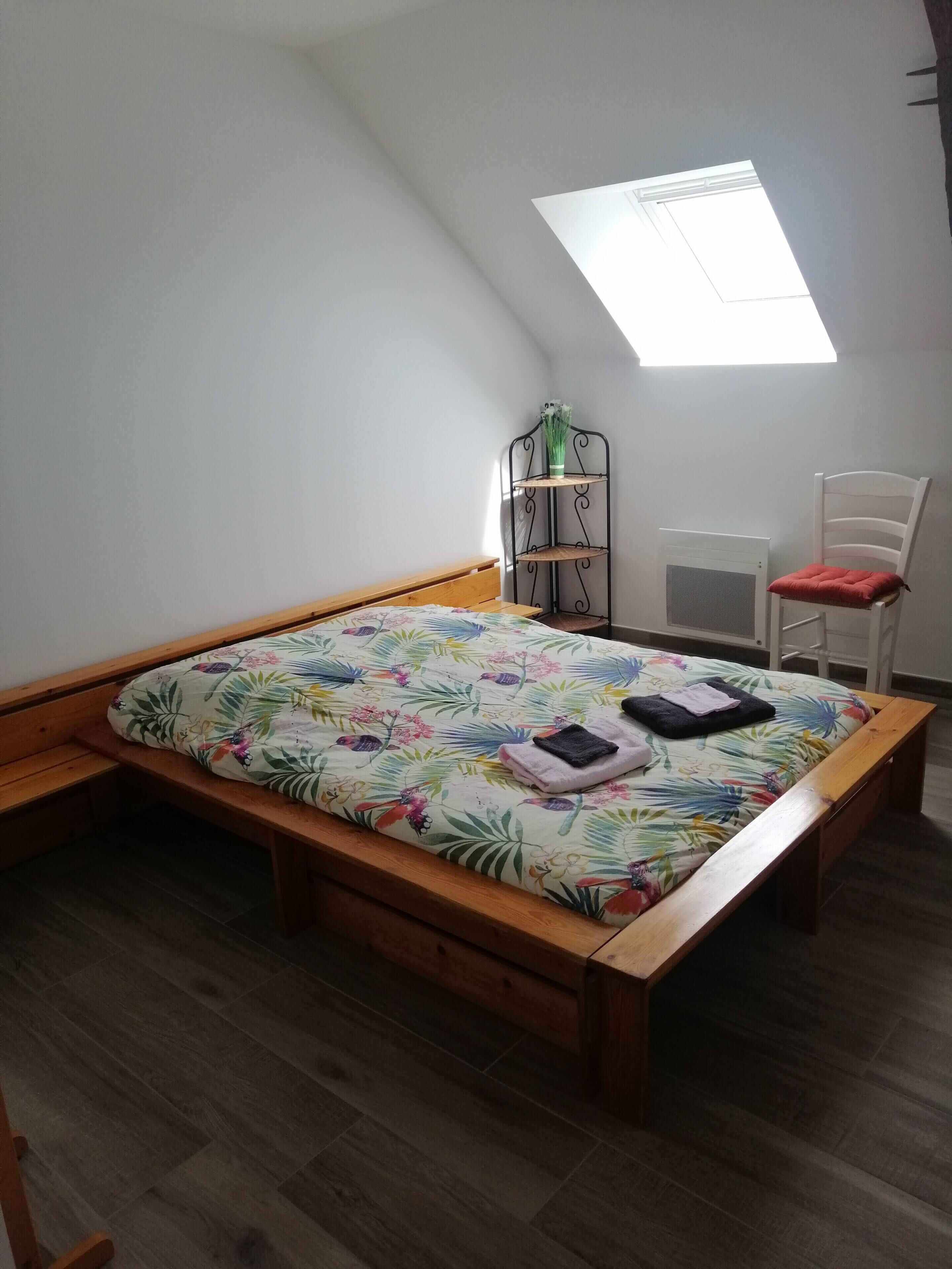 Un dormitorio con techo inclinado, una cama de madera, una pequeña silla blanca, una estantería metálica y un tragaluz.