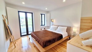 A bedroom with a large bed, wooden flooring, a bench, a small table with a lamp, and a view through a sliding door. - Duplex com Jacuzzi, Terraço, & Bbq. Vista Serra (Covilhã)