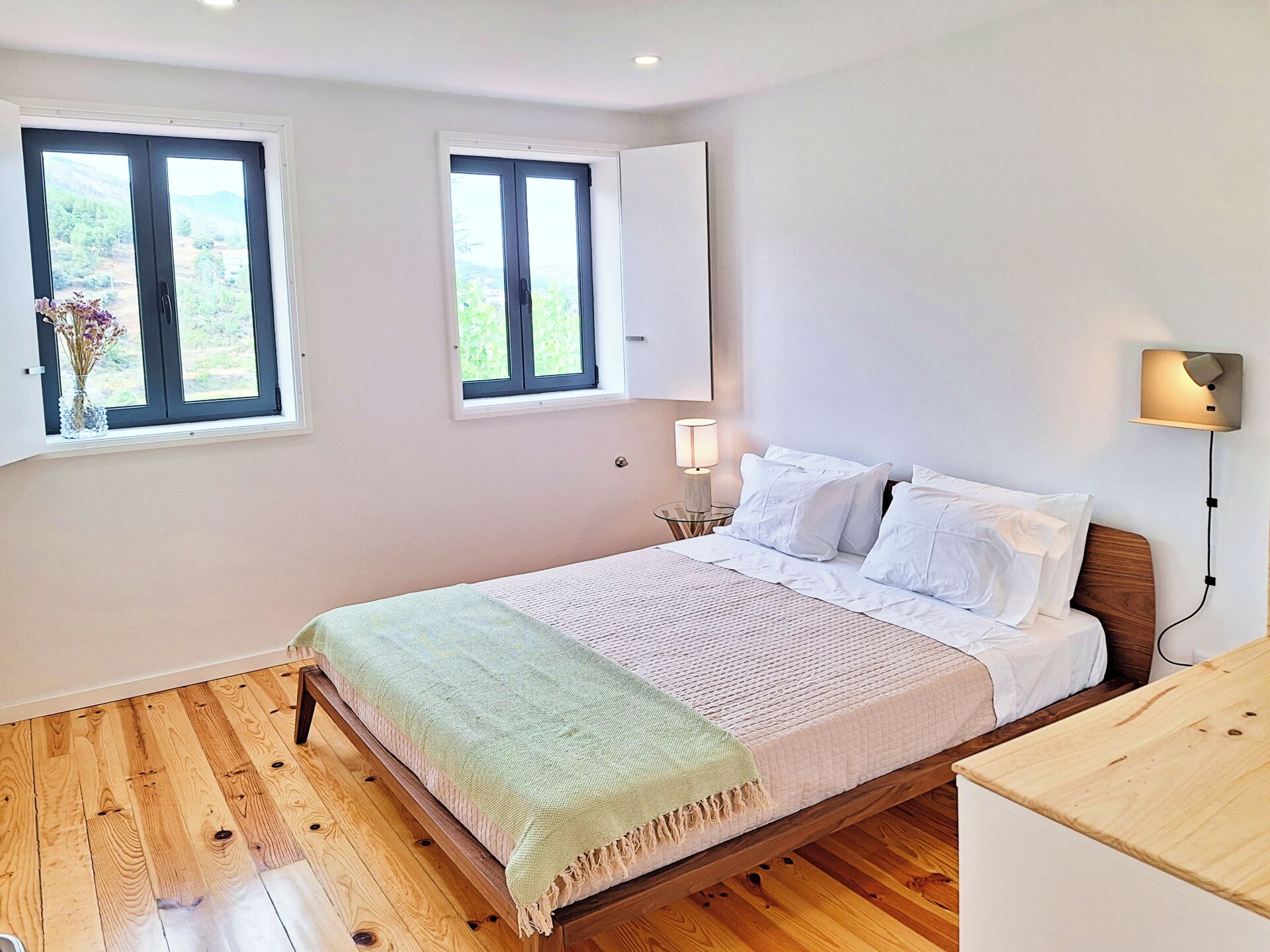 Une chambre à coucher avec un lit en bois, deux fenêtres donnant sur la verdure, une lampe de chevet et un luminaire fixé au mur.