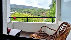 A balcony with a wicker chair and a table, overlooking a mountainous landscape with greenery and distant buildings. - Duplex com Jacuzzi, Terraço, & Bbq. Vista Serra (Covilhã)