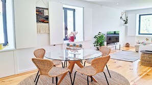 A modern dining area with a glass table surrounded by wicker chairs, a large woven rug, and a view of a kitchen and living area. - Duplex com Jacuzzi, Terraço, & Bbq. Vista Serra (Covilhã)