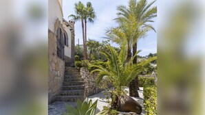 Un escalier en pierre mène à un bâtiment blanc, surmonté d’une grille en métal, et l’ensemble est entouré de palmiers et d’un jardin agrémenté d’une grande plante et d’un pot décoratif.