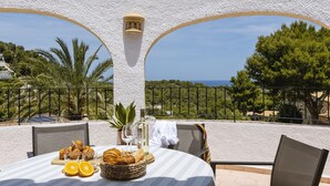 Une table dressée avec du pain, des oranges et des verres à vin, offrant une vue panoramique sur un paysage pittoresque, avec des arbres et la mer à l'horizon.