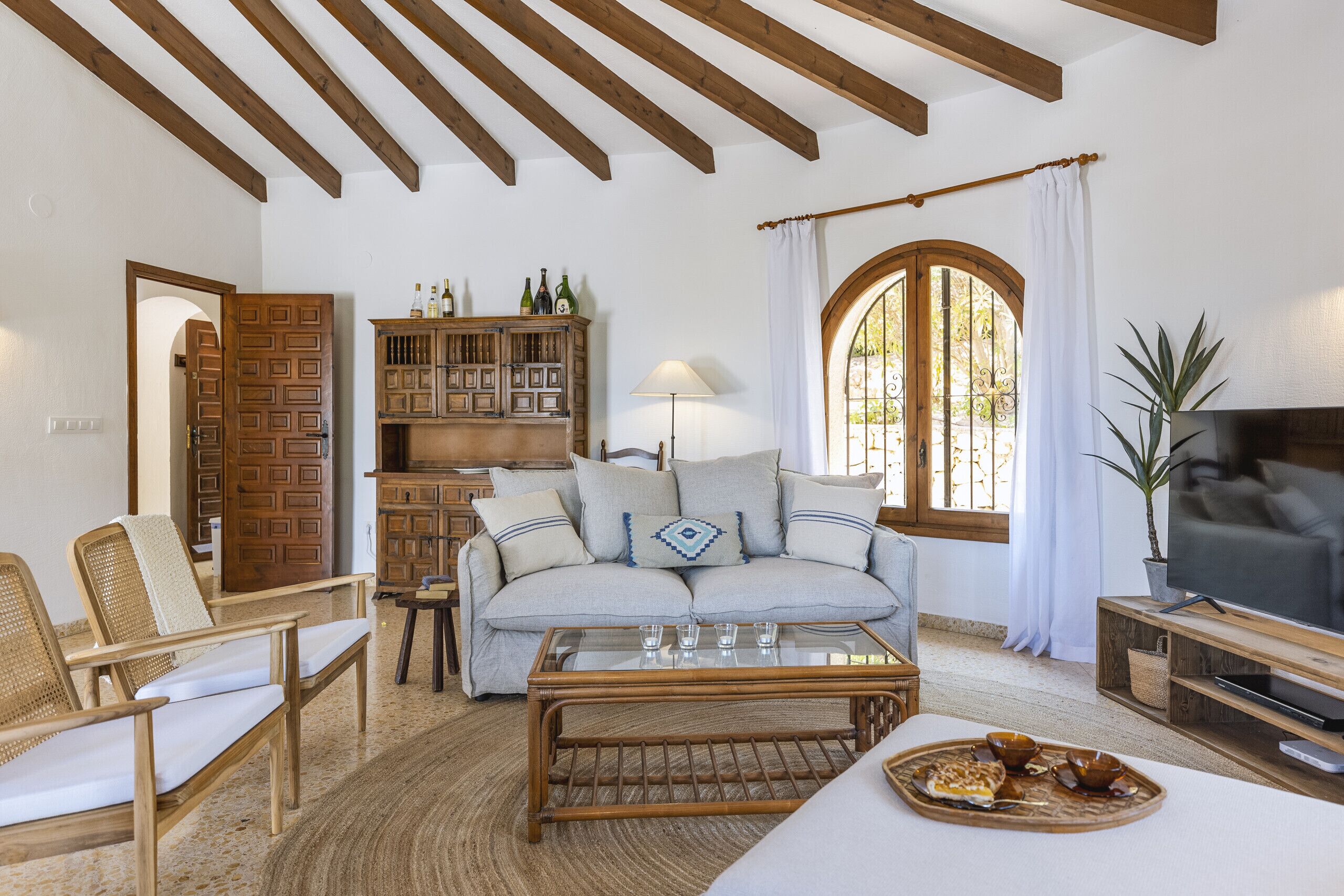 A cozy living room with a sofa, chairs, a coffee table, and a TV. There's a bookshelf and a window with curtains.