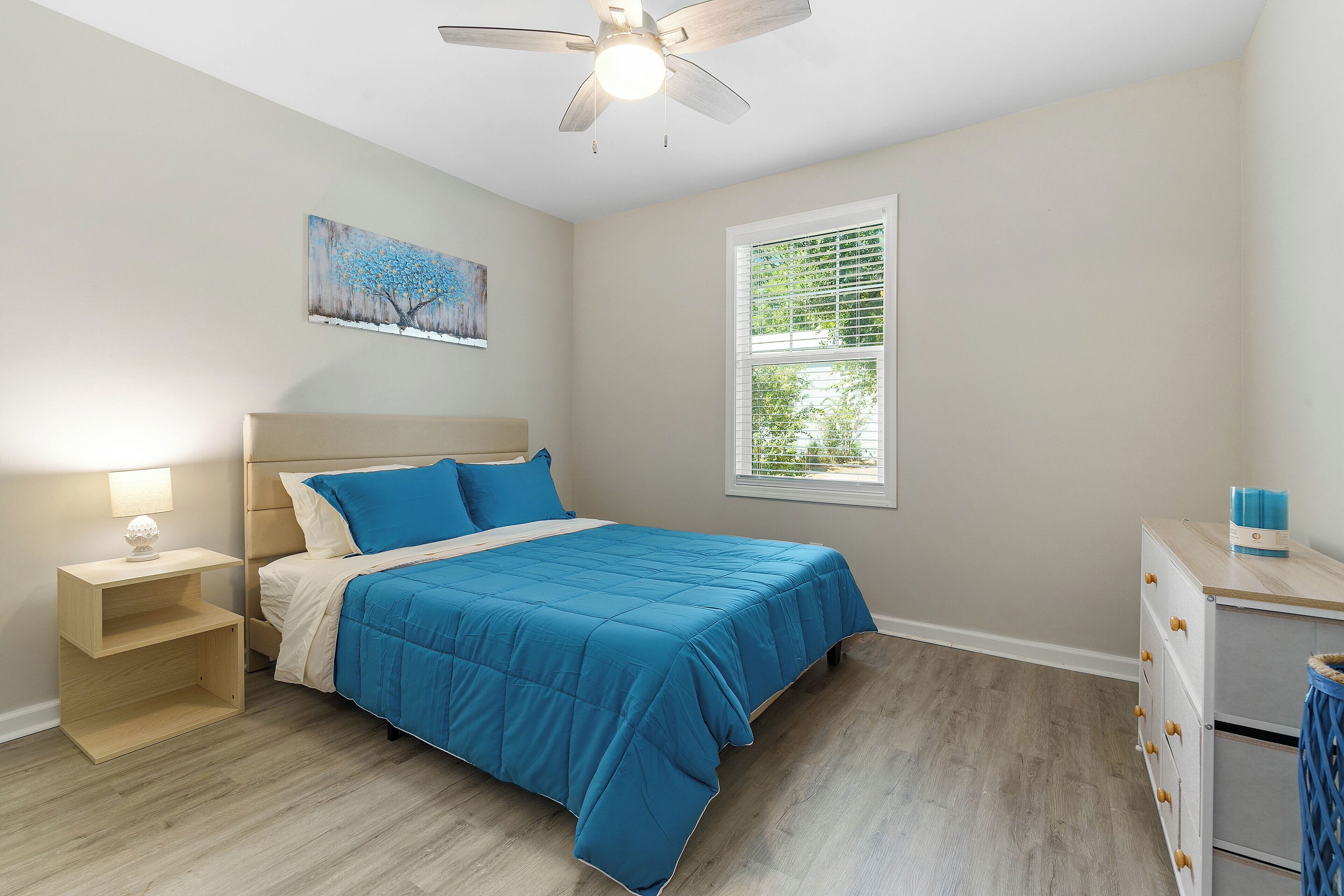 A bedroom with a bed, a nightstand, a dresser, a ceiling fan, a window, and a painting on the wall.