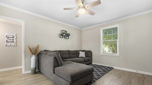 A living room with a sectional sofa, a ceiling fan, a window with blinds, and a decorative wall piece. - The Tulip in Midtown (Mobile)