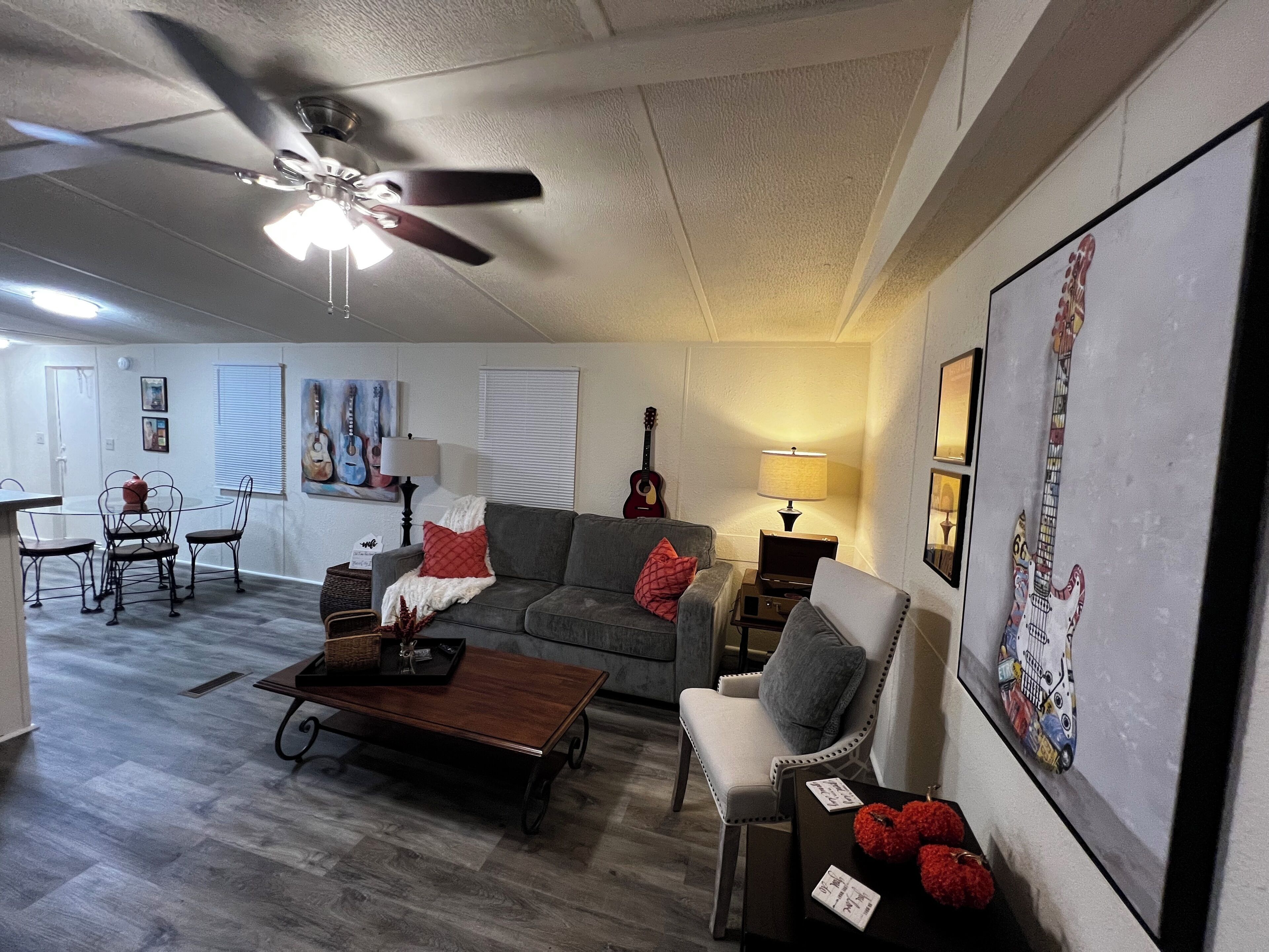 A living room with a ceiling fan, a dining area with a table and chairs, a gray sofa with decorative pillows, a wooden coffee table, and a wall with framed artwork.