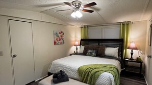 A bedroom with a bed, a bench, a ceiling fan, a nightstand with a lamp, and a door.