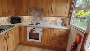 A compact kitchen with wooden cabinets, a microwave, a stove, and a sink. - Self-catering holiday Chalet in Cornwall (Saint Merryn)