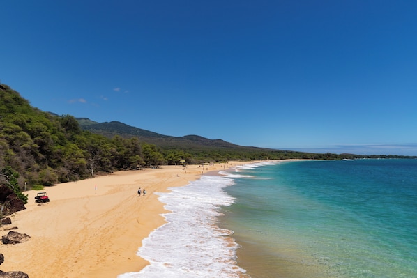 Une plage aux eaux turquoise et limpides, bordée d’un rivage sablonneux, avec une chaîne de montagnes en toile de fond.