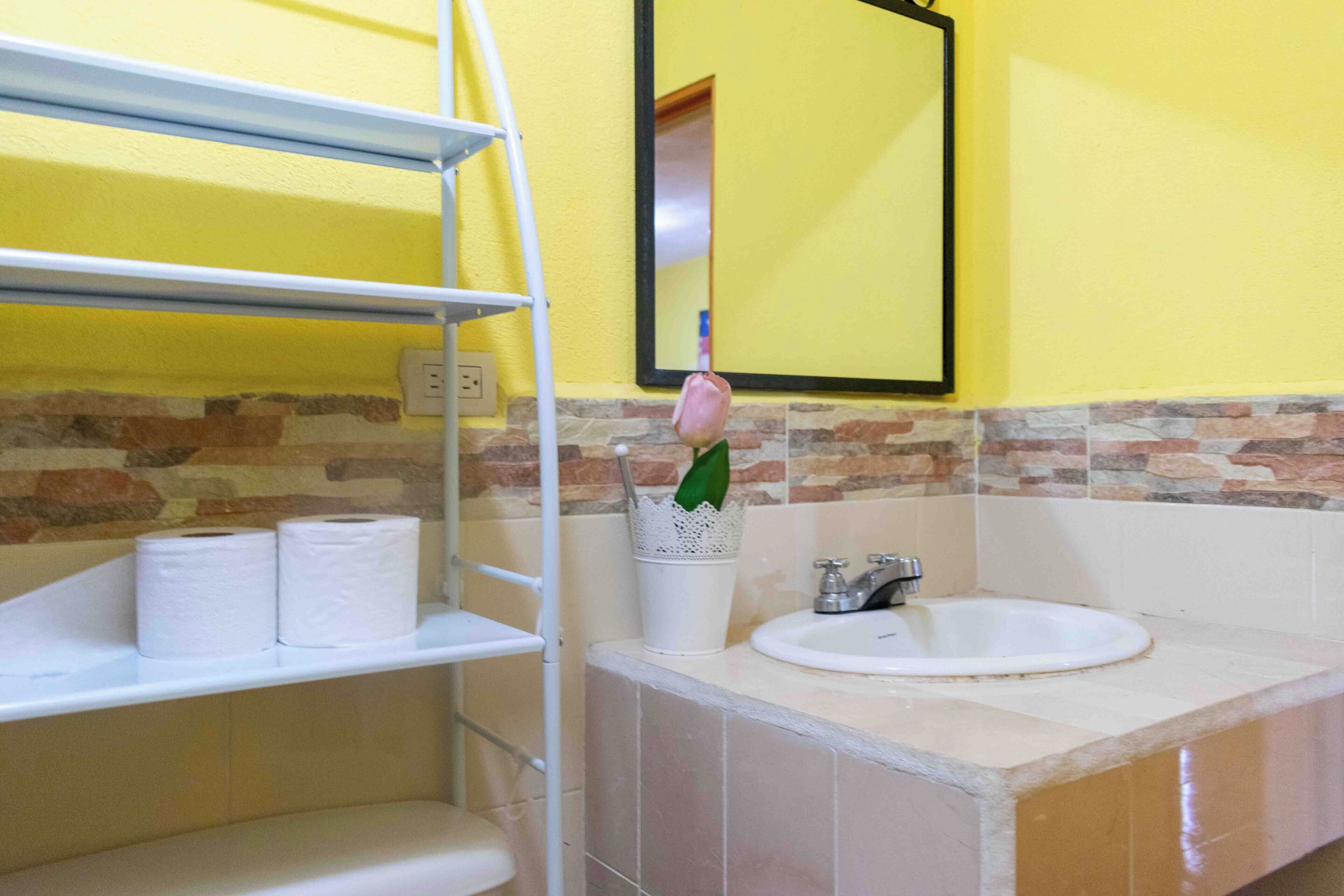 A bathroom with a yellow wall, a sink, and a mirror. There are two rolls of toilet paper on a shelf and a white cup with a green plant.