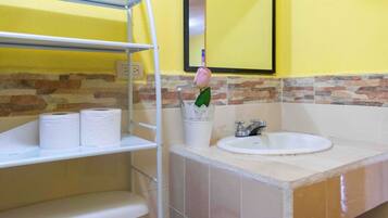 A bathroom with a yellow wall, a sink, and a mirror. There are two rolls of toilet paper on a shelf and a white cup with a green plant.