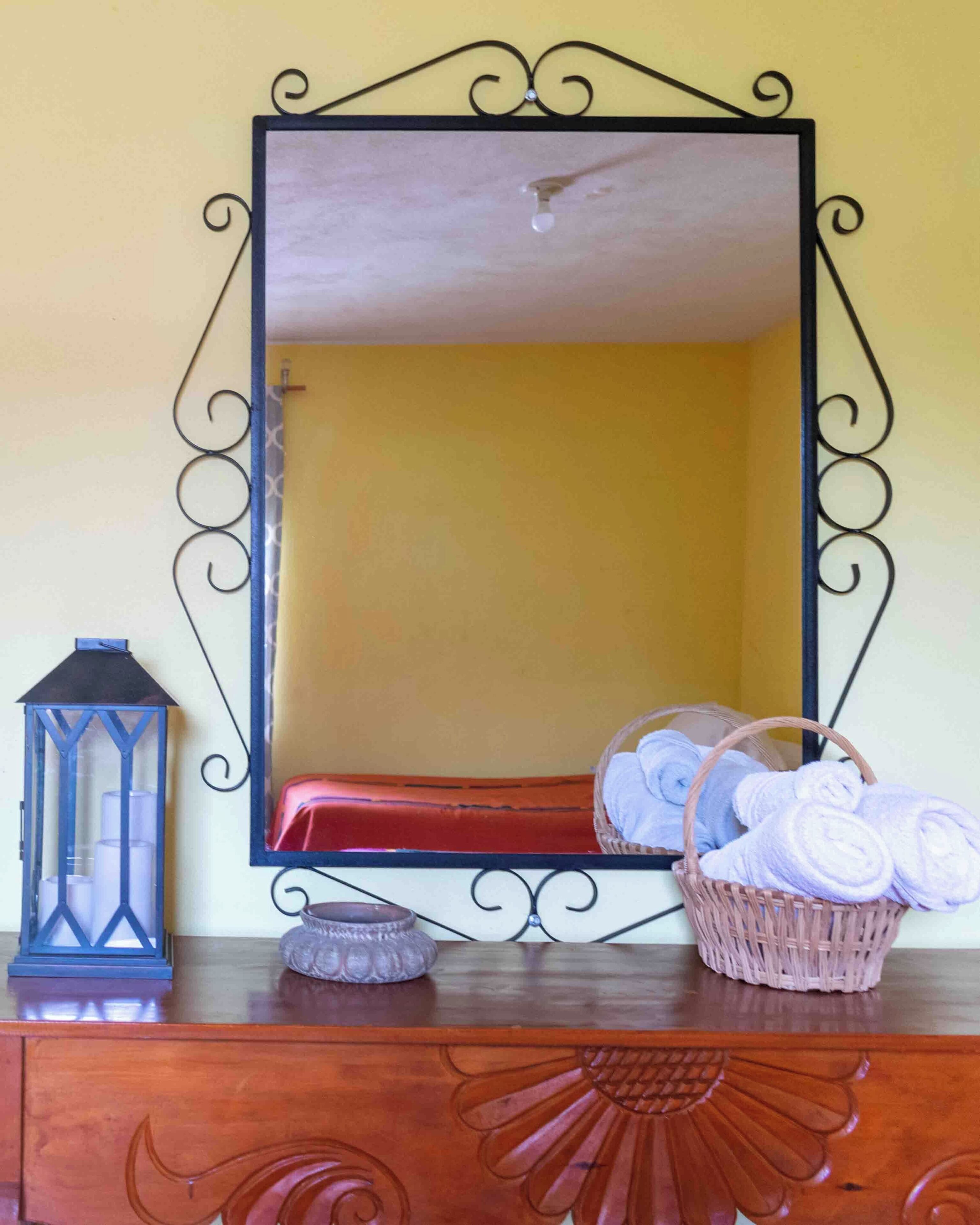 Un espejo con marco negro decorativo, un aparador de madera con espejo, un farol azul y una cesta de toallas enrolladas.
