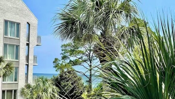Um edifício com vários andares e varandas, rodeado por plantas tropicais e vista clara para o oceano.
