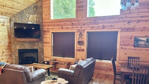 A cozy cabin interior with a stone fireplace, leather sofas, a flat-screen TV, and large windows offering a view of greenery.