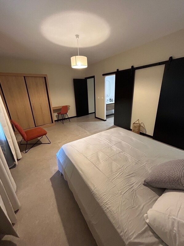 Un dormitorio con una cama grande, una silla, un armario y un baño visible a través de una puerta abierta.