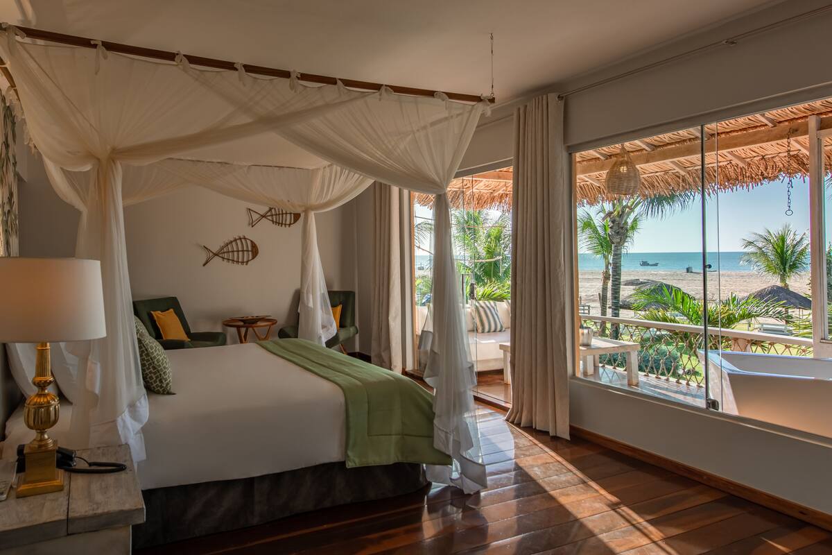 Un dormitorio con cama con dosel, vista a la playa y un balcón con techo de paja.