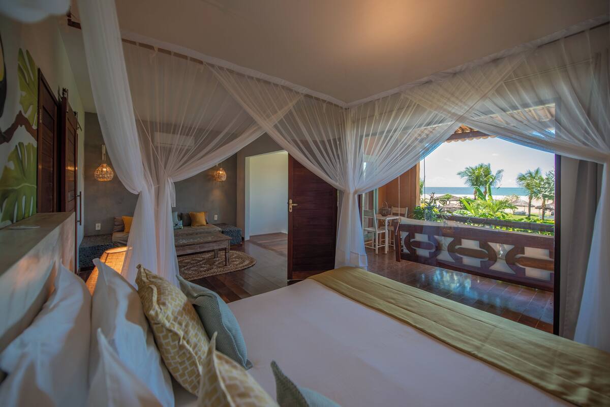 Un dormitorio con una cama grande, un balcón con vista a la playa y una zona de comedor con una mesa y sillas.