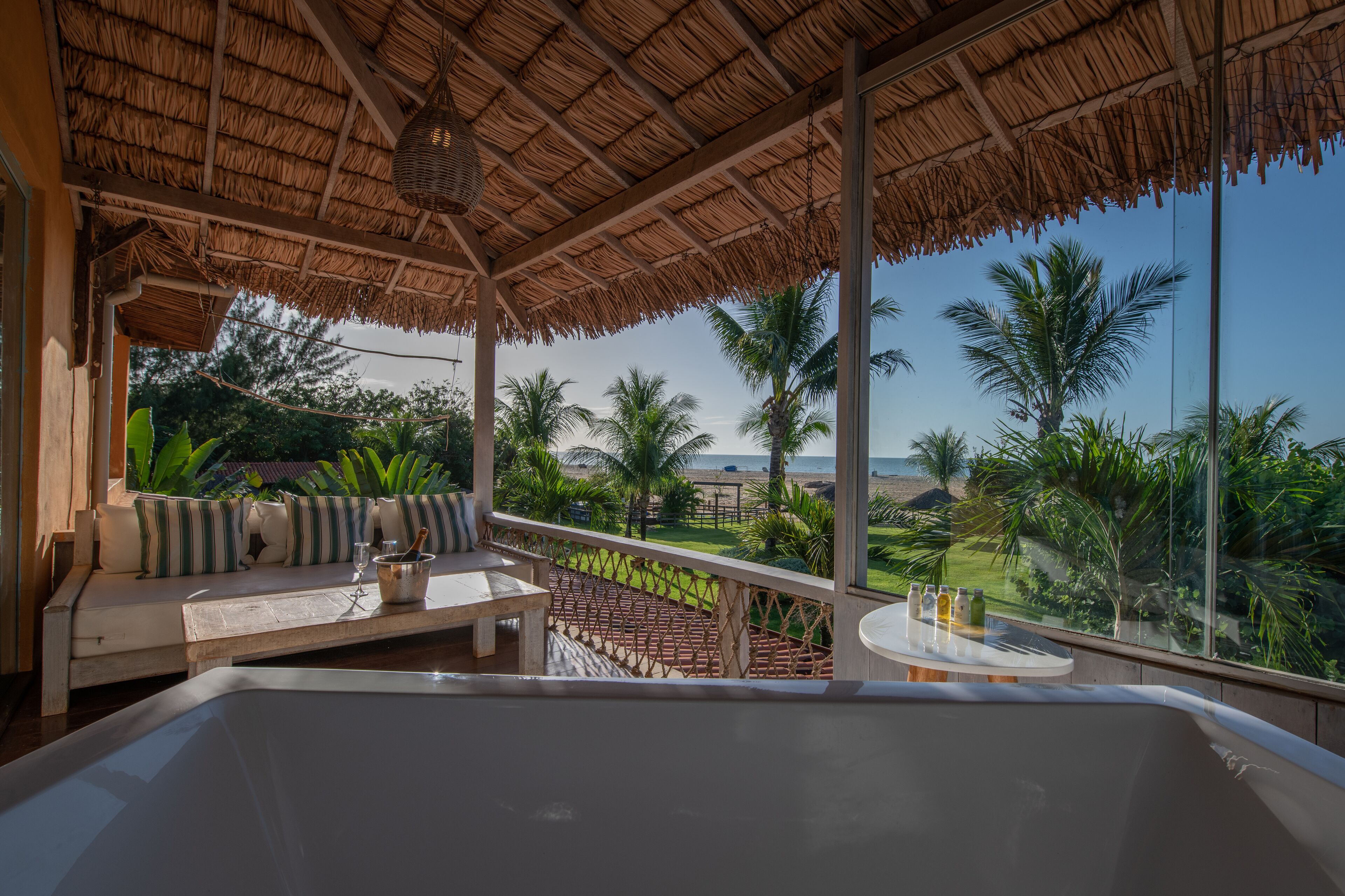 A balcony with a thatched roof, a seating area with cushions, and a view of the beach and palm trees.
