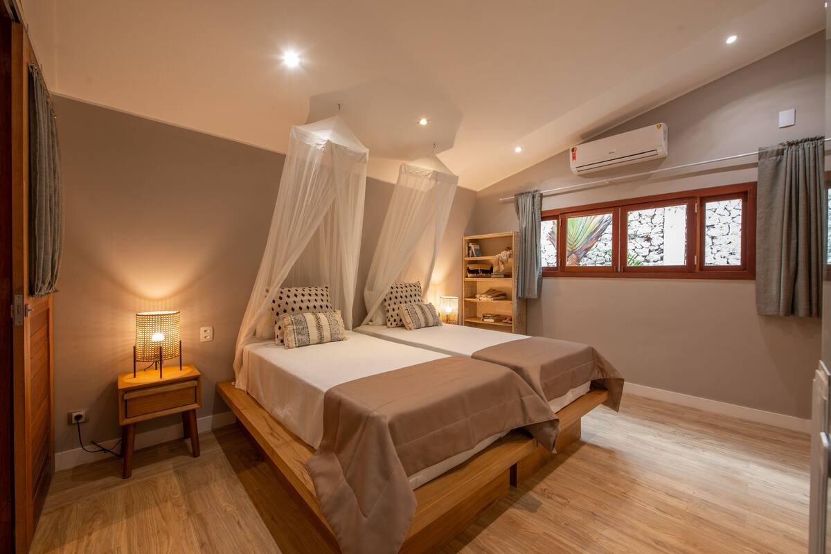Un dormitorio con dos camas, una mesita de noche, una estantería con libros y una ventana con cortinas.