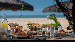 Ein Frühstückstisch mit Speisen und Getränken, mit Blick auf einen Strand und Strandstühle mit Reetdach.