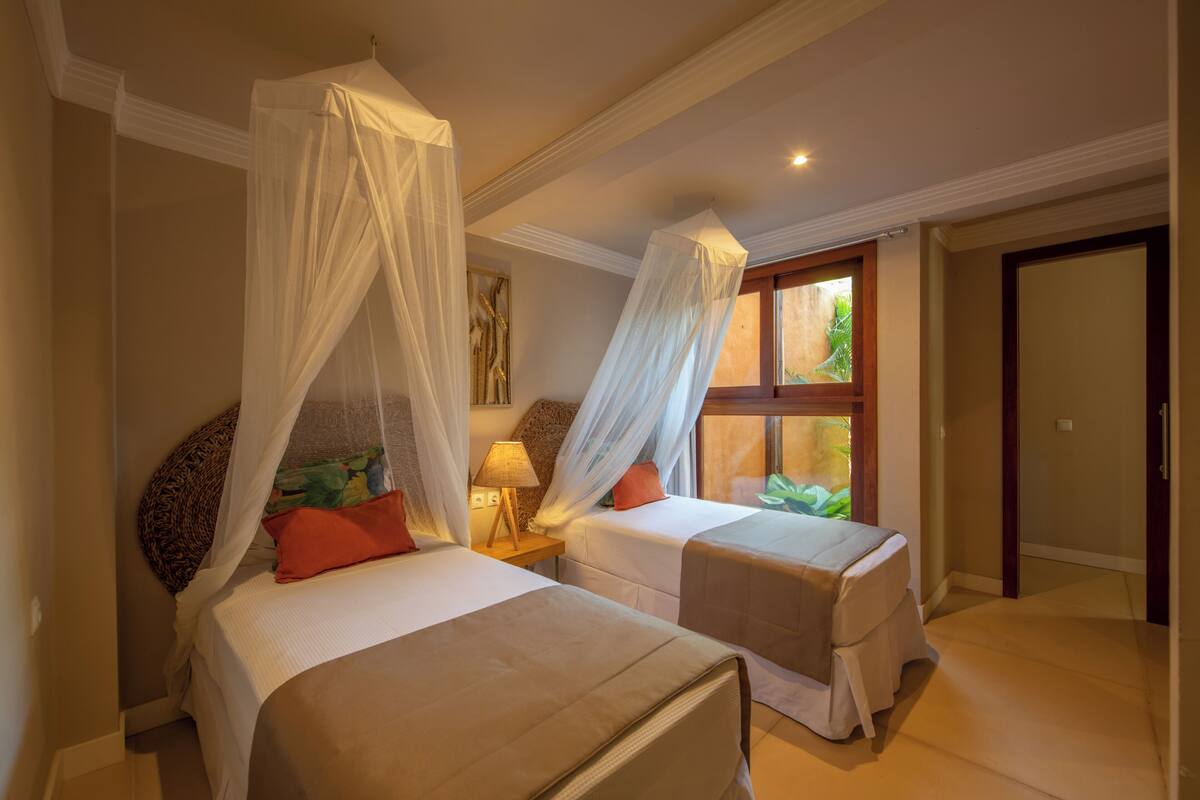 Un dormitorio con dos camas, cada una con dosel, una mesita de noche con lámpara y una ventana con vistas a follaje.