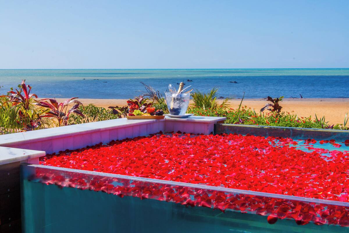 Una piscina con pétalos de flores rojas, una copa de champán y vistas a la playa y al océano.