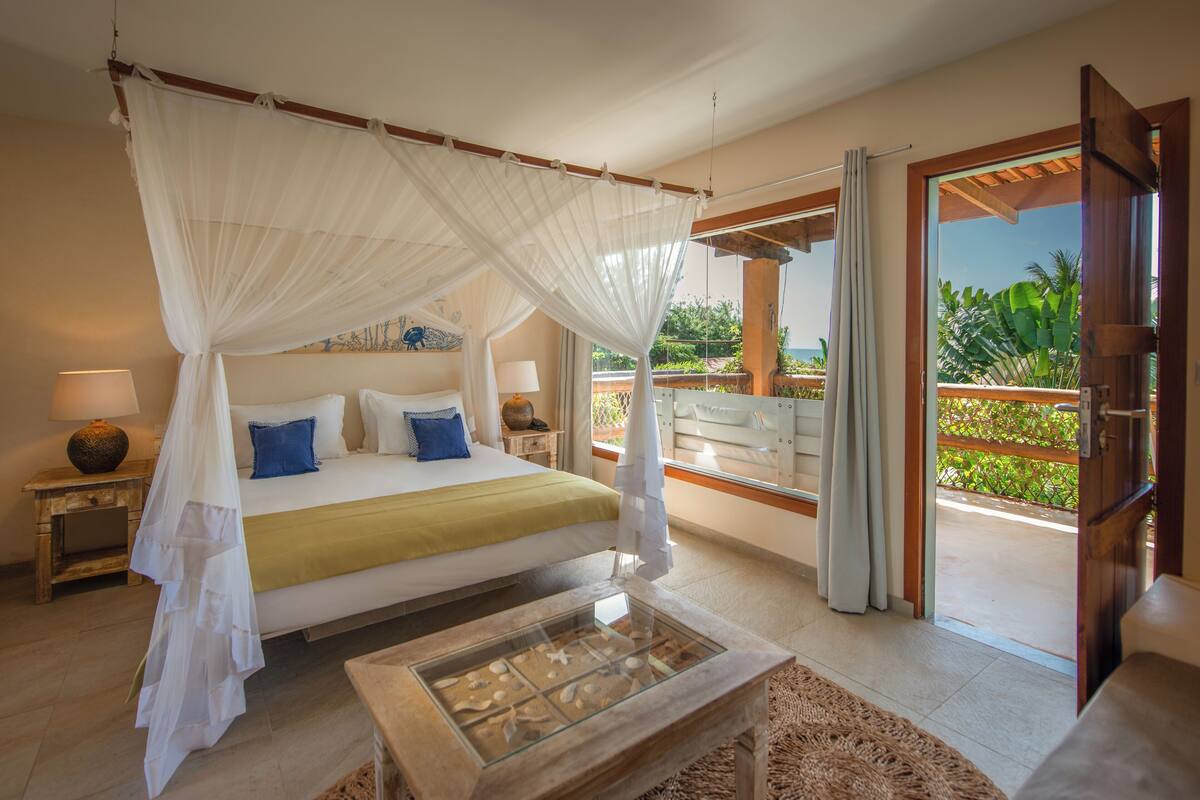 Un dormitorio con cama con dosel, una mesita de centro de vidrio y objetos decorativos, y vistas al exterior.