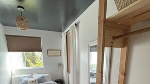A bedroom with a bed, a window with blinds, a bedside table, and a view of the outdoors. - Fireweed Hollow Junior - Cozy and cute modern cabin (Homer)