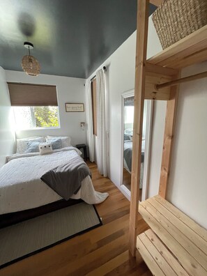 A bedroom with a bed, a window with blinds, a bedside table, and a view of the outdoors. - Fireweed Hollow Junior - Cozy and cute modern cabin (Homer)