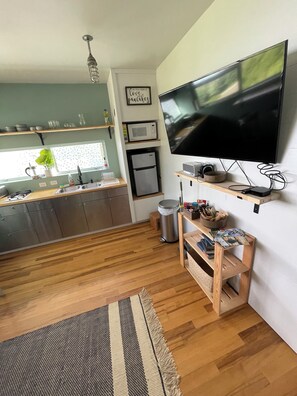 A modern kitchen with a flat-screen TV, wooden shelves, a microwave, and a refrigerator. - Fireweed Hollow Junior - Cozy and cute modern cabin (Homer)