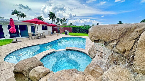 A swimming pool with a rock waterfall feature, surrounded by lounge chairs and a red umbrella. - Waterfront Oasis – Private Heated Pool & Waterfall Hot Tub (Pompano Beach)
