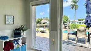 A room with a sliding glass door leading to an outdoor area with a pool, lounge chairs, and umbrellas. - Waterfront Oasis – Private Heated Pool & Waterfall Hot Tub (Pompano Beach)