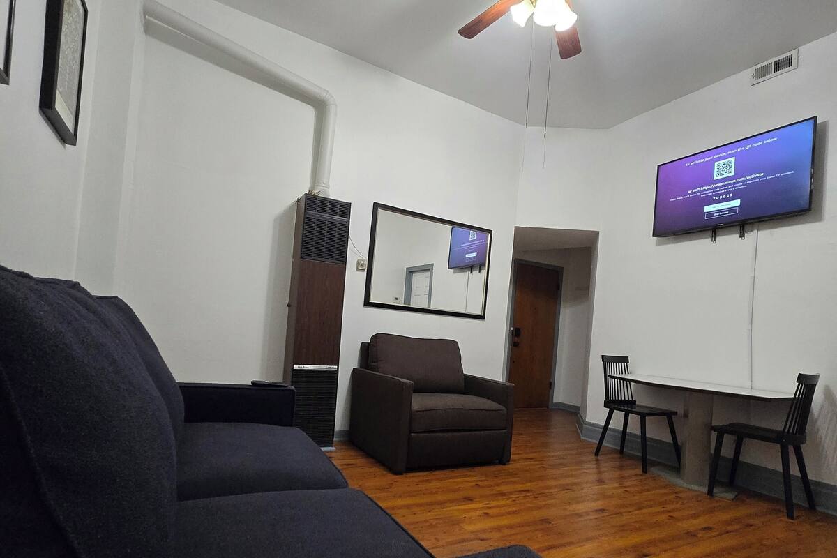 Ein Wohnzimmer mit Deckenventilator, einem an der Wand montierten Fernseher, einer Couch, einem Sessel und einem kleinen Tisch.
