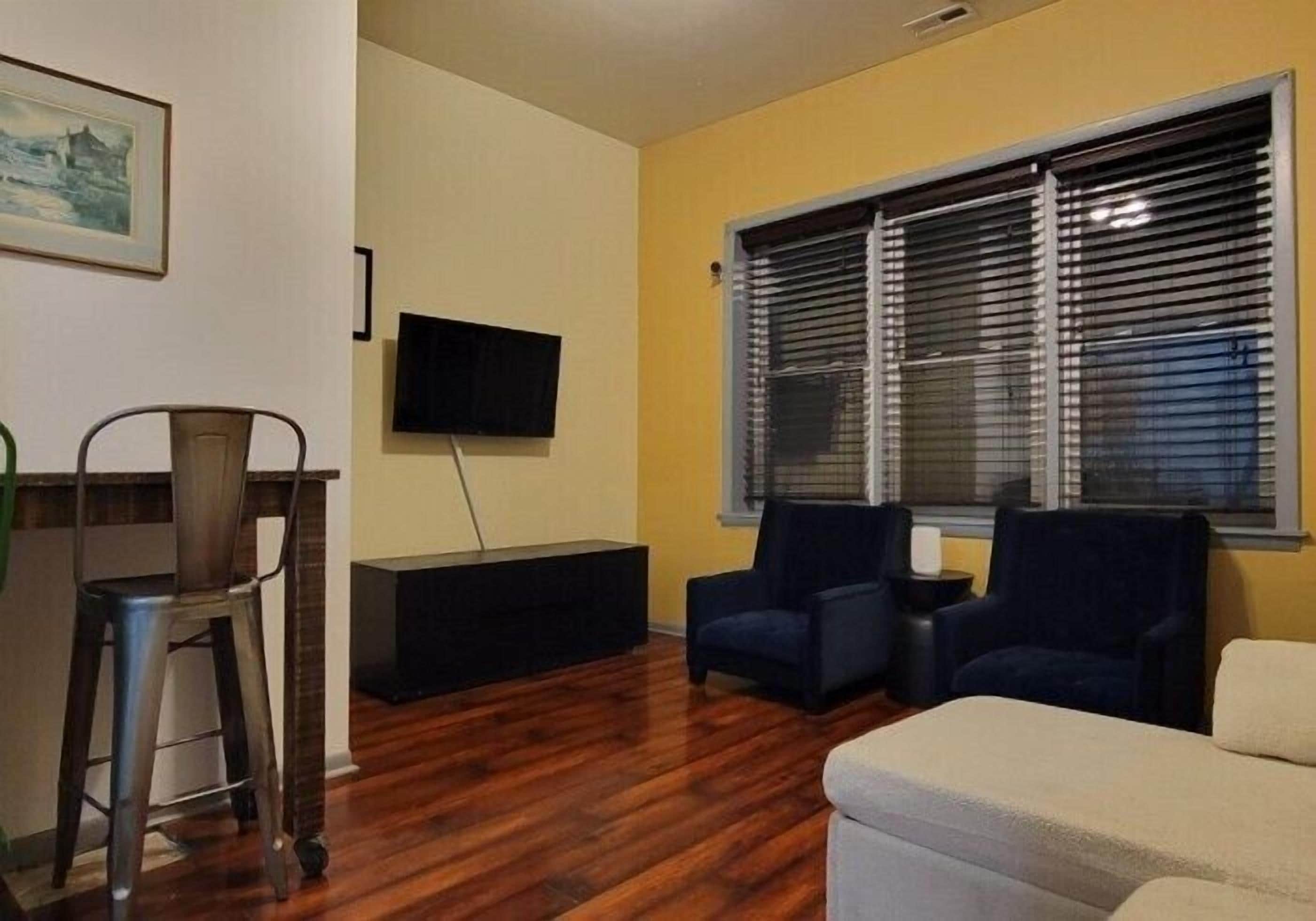 Una sala de estar con televisión, dos sillones, un sofá, una mesa de comedor con sillas y una ventana con persianas.