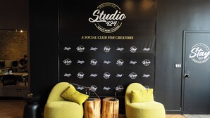 A modern interior with a black wall featuring the 'Studio 124' logo and text. There are two yellow chairs with wooden stools in front, a black trash bin, and a door with the same logo. - STAY (Chicago)