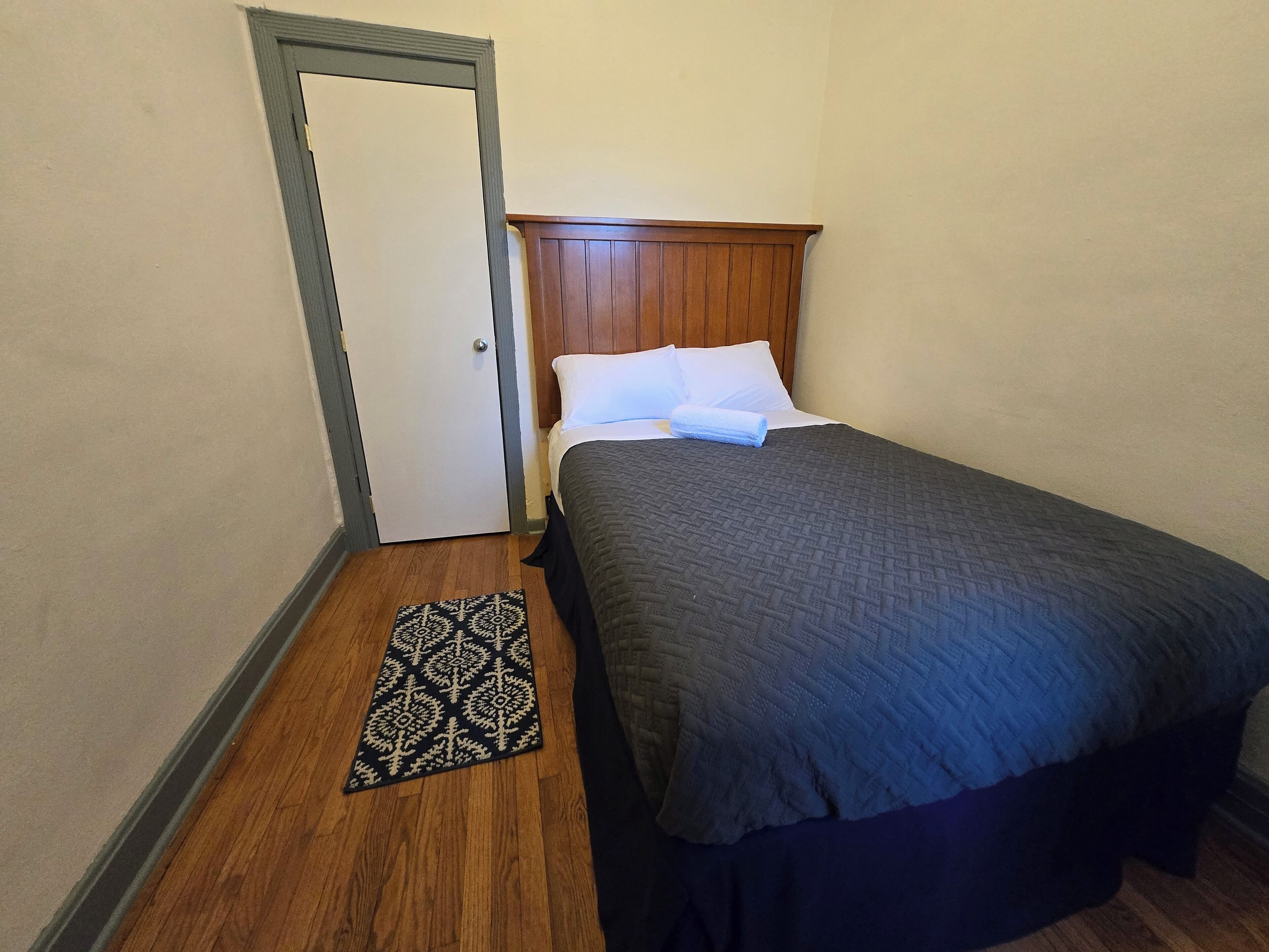 Quarto com cabeceira de madeira, cama com colcha escura, travesseiro branco e tapete estampado no chão.